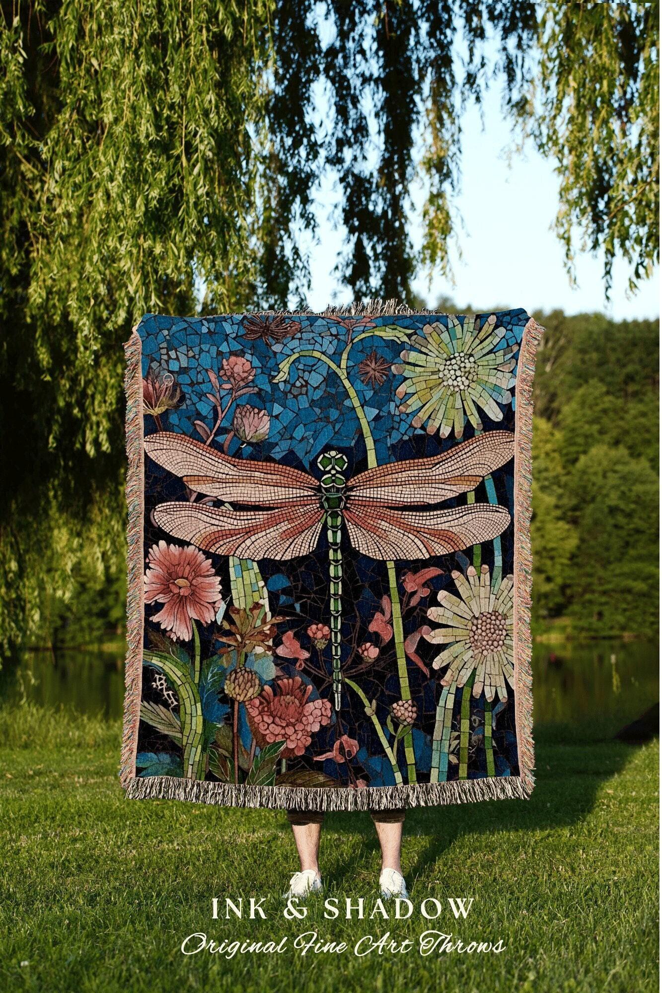 May include: A decorative throw blanket featuring a large dragonfly design with a mosaic-style background. The dragonfly has pink and brown wings, a green body, and is surrounded by flowers in shades of pink and green. The blanket has a fringed edge.