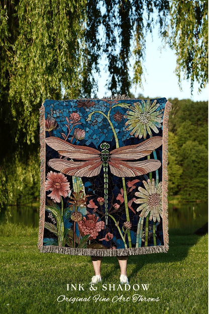 May include: A decorative throw blanket featuring a large dragonfly design with a mosaic-style background. The dragonfly has pink and brown wings, a green body, and is surrounded by flowers in shades of pink and green. The blanket has a fringed edge.