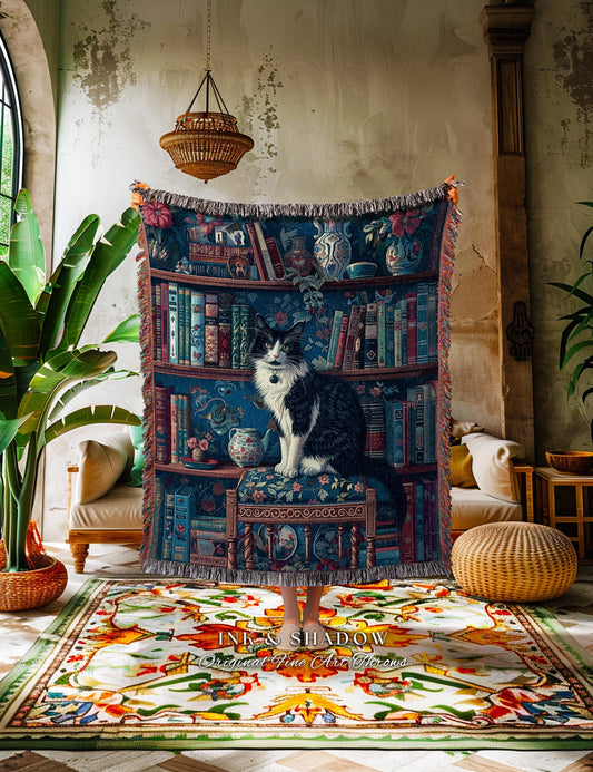 May include: A tapestry throw blanket featuring a bookshelf design with a black and white cat perched on a floral patterned seat. The throw has a blue background with books, vases, and flowers. The blanket has a fringed edge.