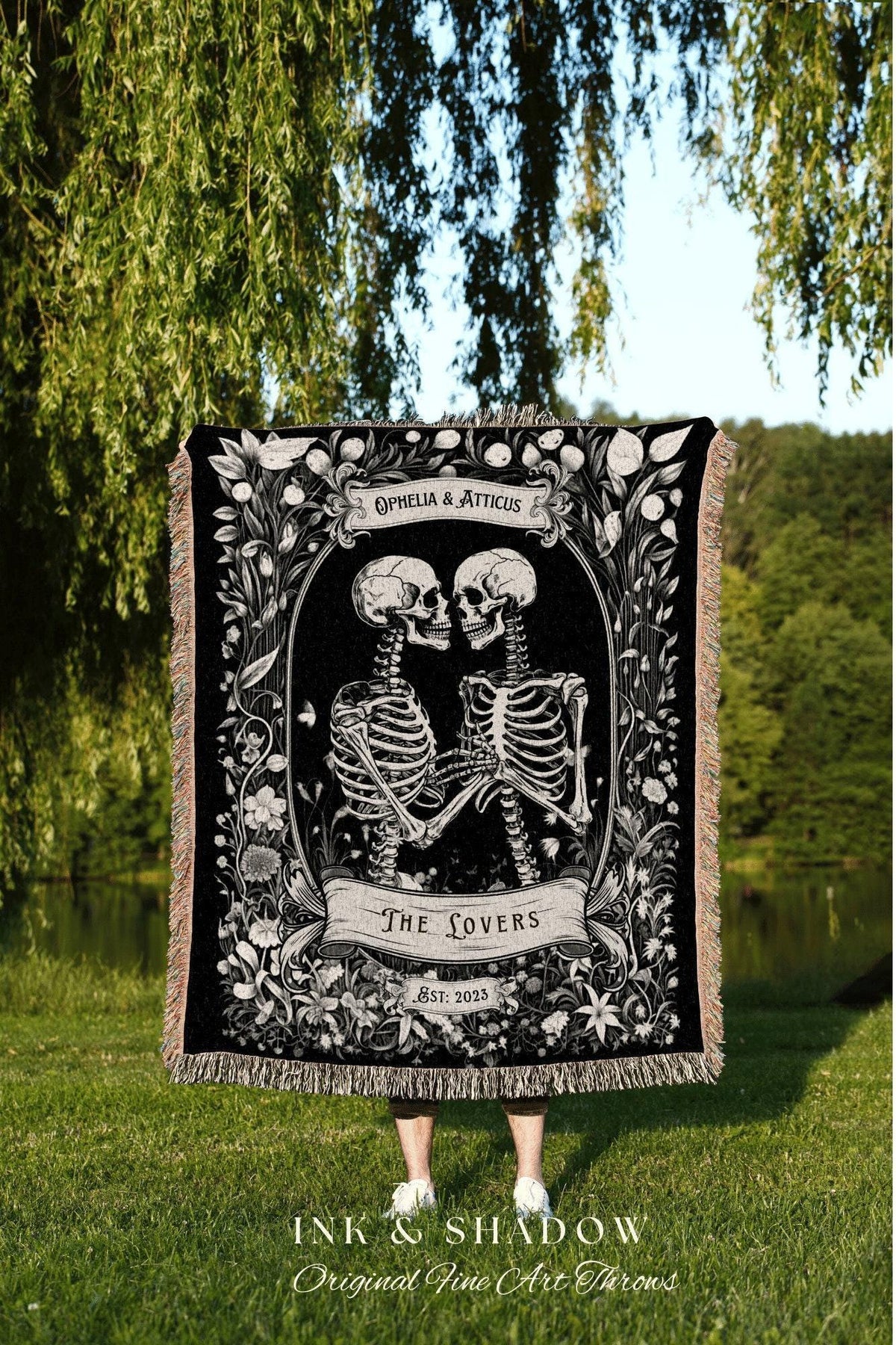May include: Black and white throw blanket featuring a skeletal couple embracing within a floral frame. The blanket reads "Ophelia & Atticus" and "The Lovers, Est. 2023." The fringe is a light tan color. The blanket is held outdoors.
