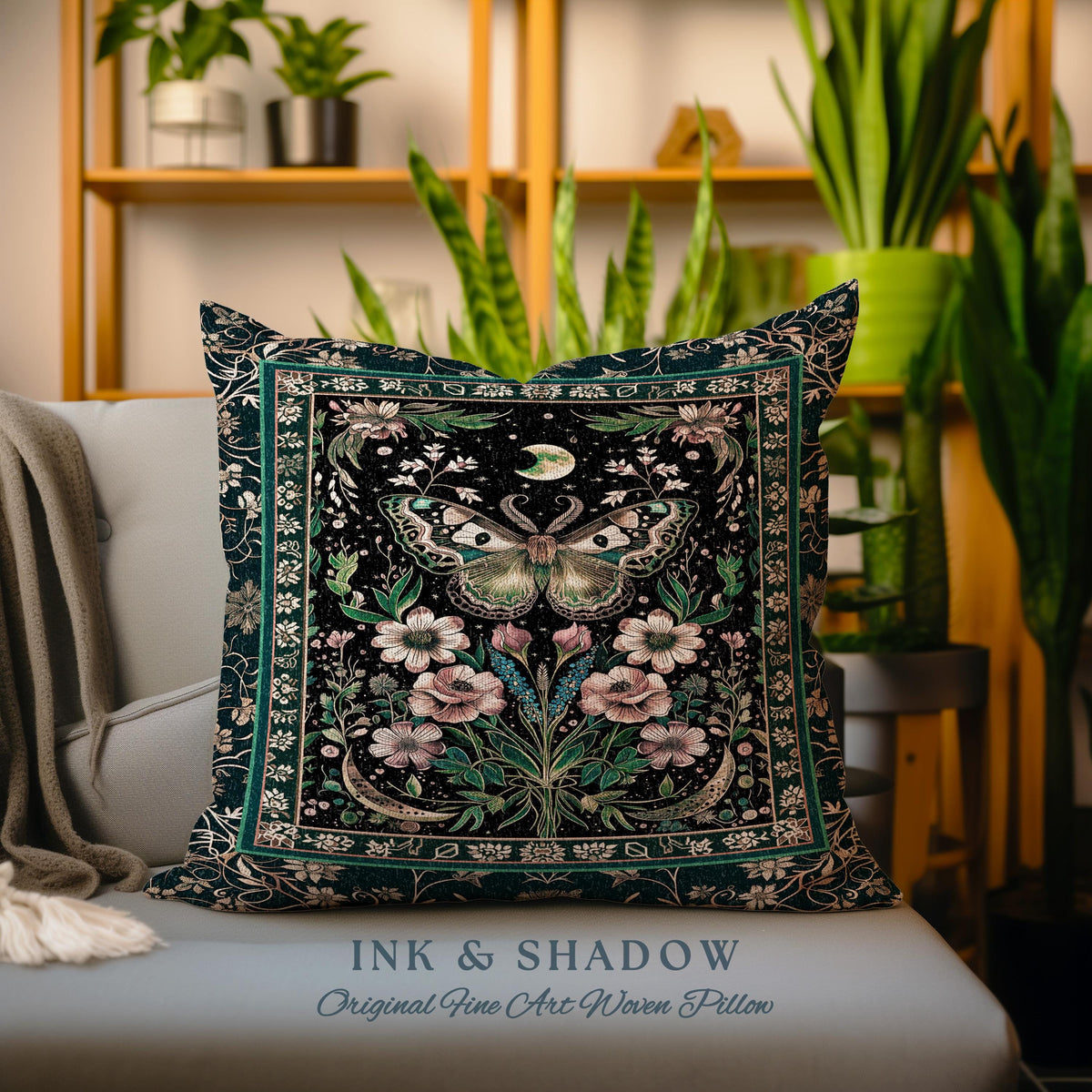 May include: A dark green throw pillow with a black background featuring a detailed illustration of a moth with pink flowers and leaves. The pillow has a gold border and the text "INK & SHADOW Original Fine Art Woven Pillow" at the bottom.