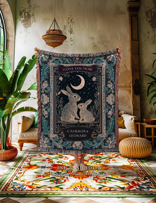 May include: A woven tapestry blanket with a dark teal background, featuring a central design of two rabbits under a crescent moon. The blanket has a decorative border and the text "I LOVE YOU MORE" and "CAMERON & LEONARD".
