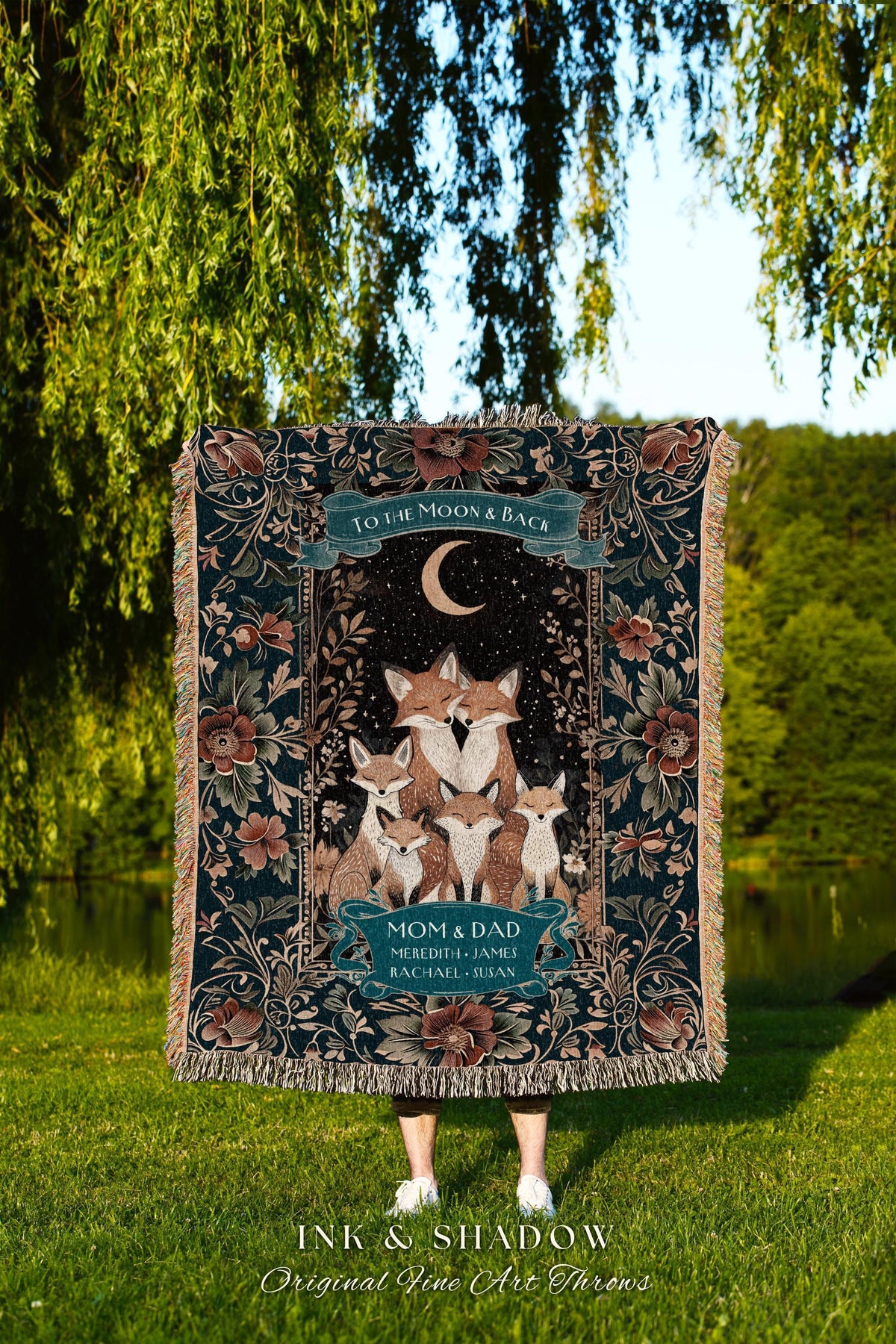 May include: A woven throw blanket with a family of foxes under a crescent moon and stars. The blanket has a dark teal background with floral accents and the text "TO THE MOON & BACK" and "MOM & DAD". The bottom of the blanket reads "INK & SHADOW Original Fine Art Throws".