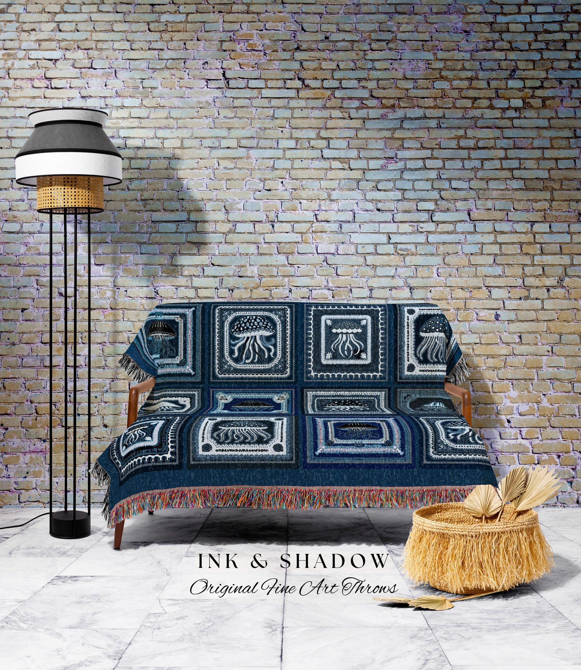 May include: A navy blue throw blanket with a jellyfish design covers a wooden sofa. The blanket has a fringed edge and is displayed in front of a brick wall. A floor lamp and a woven basket are also in the image. The text "INK & SHADOW Original Fine Art Throws" is visible.