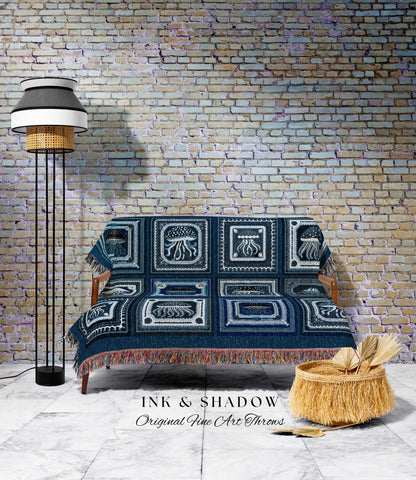 May include: A navy blue throw blanket with a jellyfish design covers a wooden sofa. The blanket has a fringed edge and is displayed in front of a brick wall. A floor lamp and a woven basket are also in the image. The text "INK & SHADOW Original Fine Art Throws" is visible.