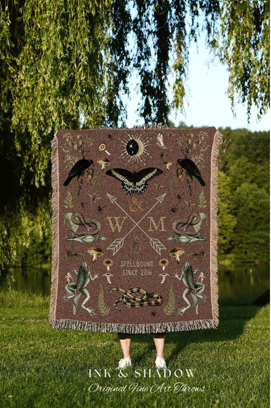 May include: A brown throw blanket with a fringe edge, featuring a detailed print of a butterfly, crows, frogs, mushrooms, and a snake. The blanket has the text "SPELLBOUND SINCE 2016" and "INK & SHADOW Original Fine Art Throws".