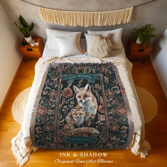 May include: A dark teal throw blanket featuring a family of foxes, surrounded by floral patterns. The blanket has a fringed edge and the text "TO THE MOON & BACK" and "FOREVER & ALWAYS". The words "INK & SHADOW" and "Original Fine Art Throws" are also visible.